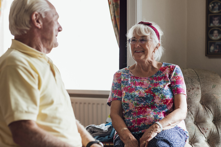 Older adult woman sitting on couch engaged in conversation with her husband.