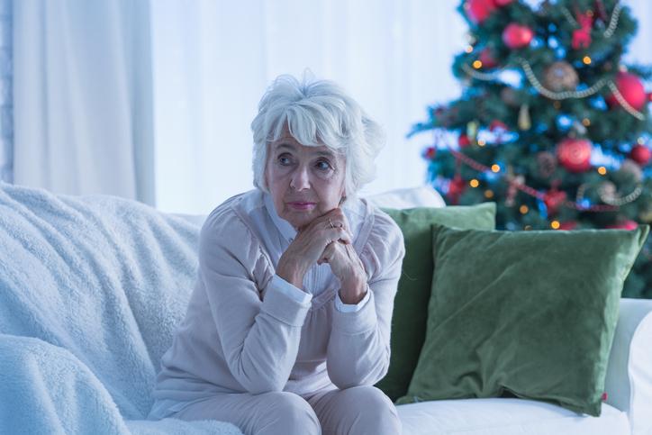 Older woman sitting alone next to Christmas tree.